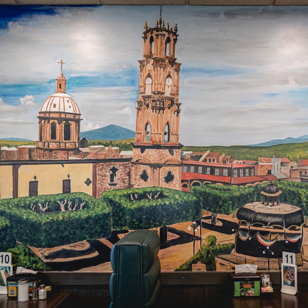 A vibrant mural of a Mexican town with a large church and clock tower, seen from a restaurant booth.