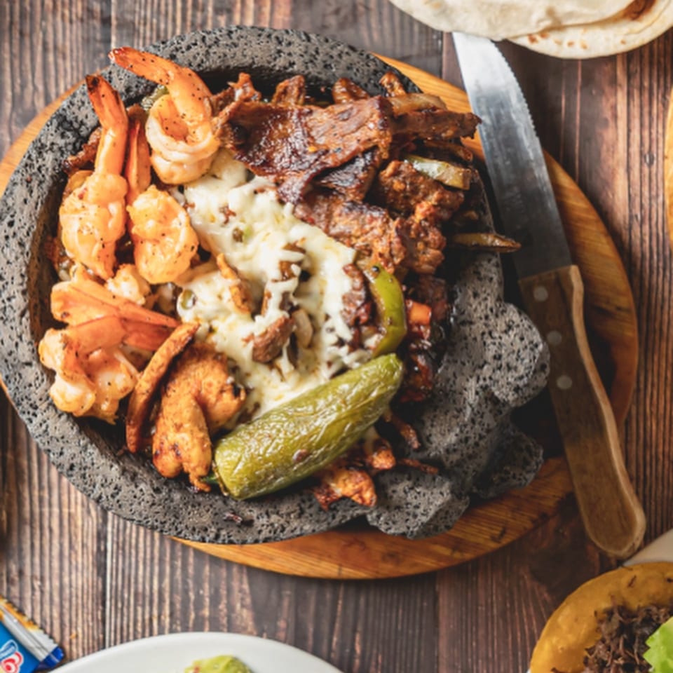 Molcajete with grilled shrimp, steak, melted cheese, and a jalapeño, served on a wooden board.