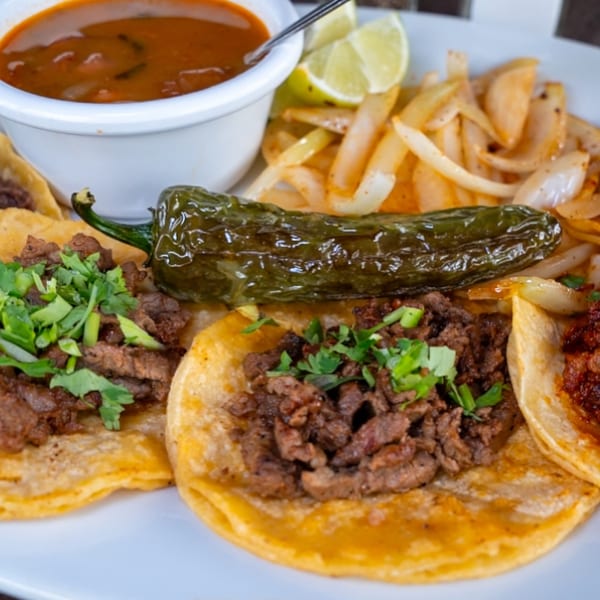 Three carne asada tacos with cilantro, a grilled jalapeño, onions, lime, and a side of consommé.
