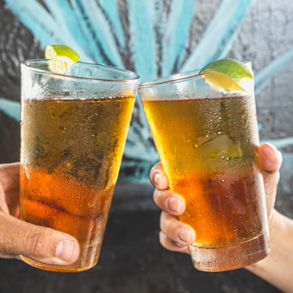 Two hands holding frosty glasses of amber liquid with lime wedges, clinking in a toast.