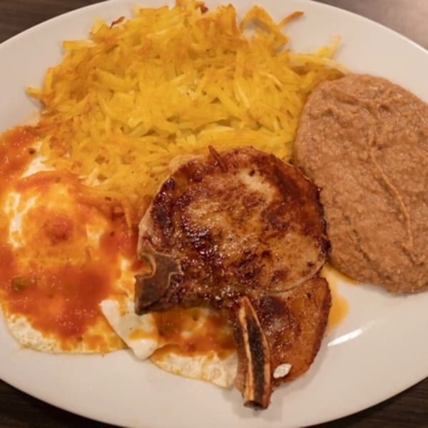 A Mexican breakfast plate with a pork chop, fried eggs with salsa, hash browns, and refried beans.