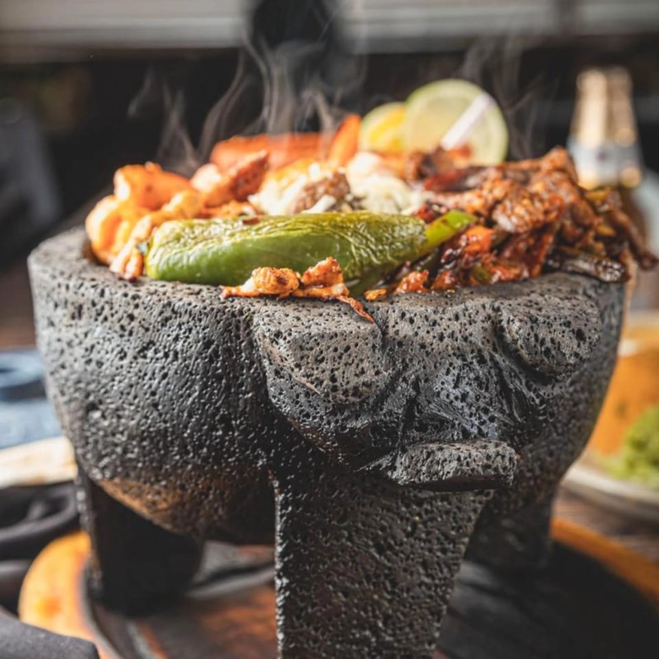 Steaming molcajete filled with shrimp, meat, cheese, and a green chili, garnished with lime.