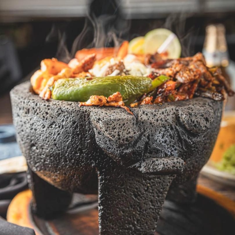Steaming molcajete filled with shrimp, meat, cheese, and a green chili, garnished with lime.