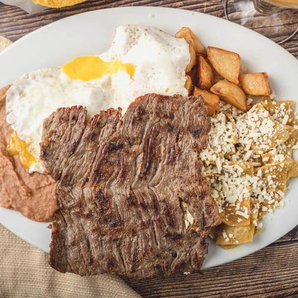 A Mexican breakfast plate with carne asada, fried eggs, refried beans, potatoes, and chilaquiles.