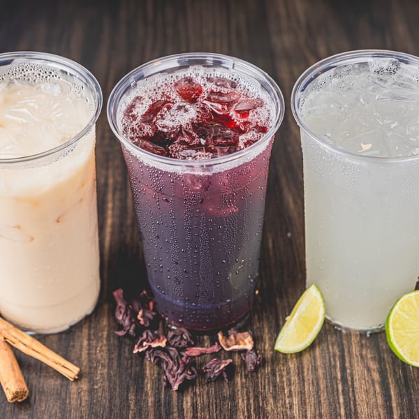 Three refreshing Mexican aguas frescas: horchata, jamaica, and limonada, with garnishes on a wooden table.