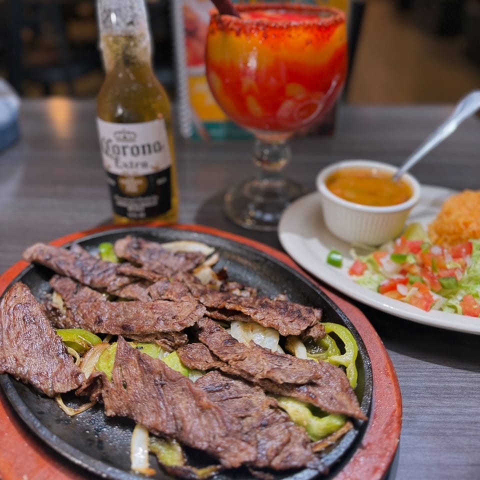 Sizzling fajitas with bell peppers and onions, served with rice, salad, a Corona, and a michelada.