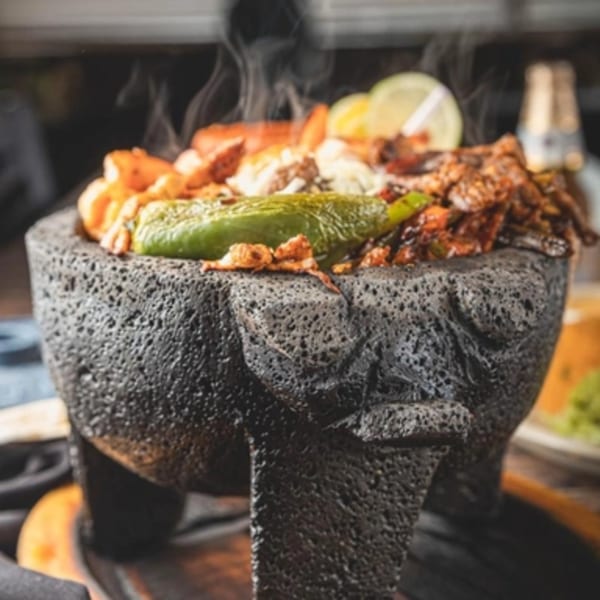 Steaming Mexican molcajete dish filled with meat, shrimp, peppers, and lime slices.