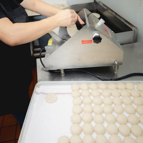 Chef's hands feeding dough into a tortilla press, with many dough balls on a tray.