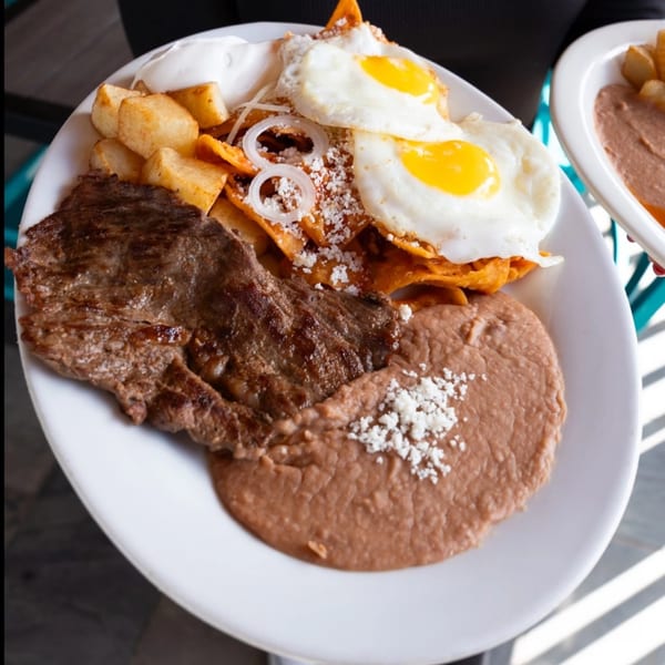 A Mexican breakfast plate with steak, refried beans, chilaquiles, fried eggs, and potatoes.