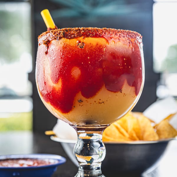 A vibrant, chili-rimmed mango margarita with a straw, salsa, and chips in the background.