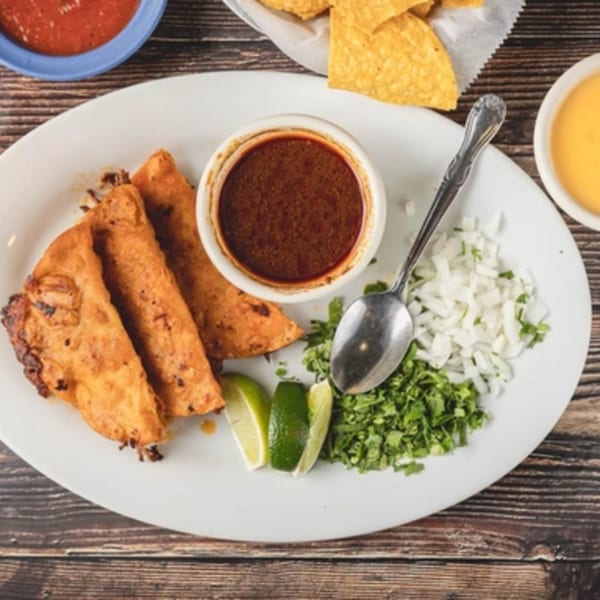 Three crispy tacos with a side of dipping sauce, cilantro, onions, and lime wedges.