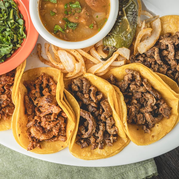 A plate of Taqueria Los Agaves tacos with various meat fillings, grilled onions, a pepper, and a side of soup.
