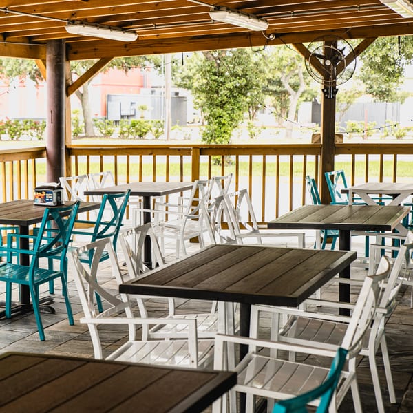 Outdoor patio with wooden ceiling, dark tables, and a mix of white and teal chairs at Taqueria Los Agaves.