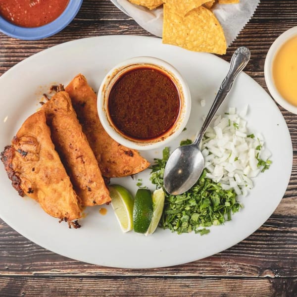 Three crispy birria tacos with consommé, cilantro, diced onions, and lime wedges on a white plate.